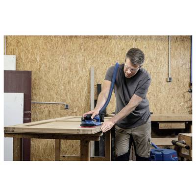 Ein Mann mit Sicherheitsbrille schleift ein Holzbrett in einer Werkstatt und verwendet dabei einen elektrischen Schleifer, der an einen Staubsauger angeschlossen ist.