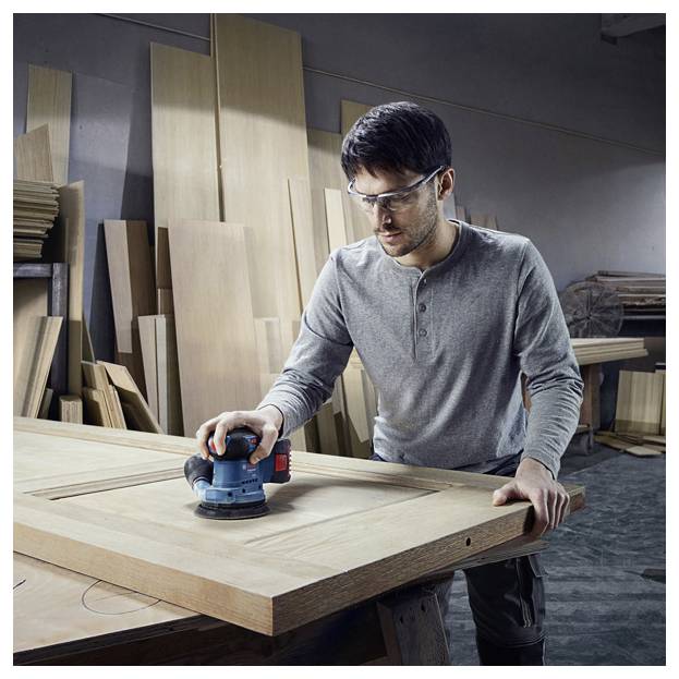 Eine Person mit Sicherheitsbrille benutzt einen elektrischen Schleifer an einer großen Holzplatte in einer Holzwerkstatt, umgeben von Holzbrettern.