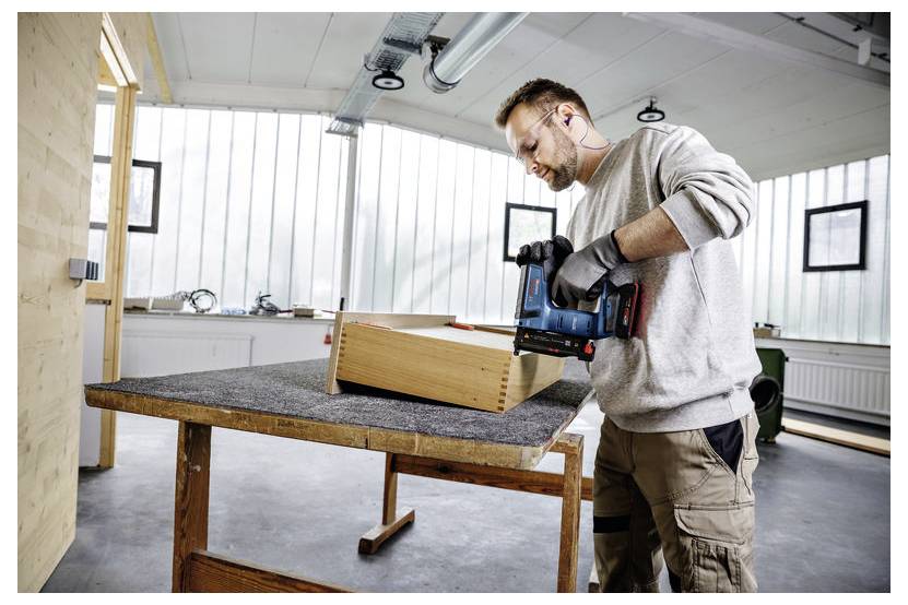 Eine Person, die ein Elektrowerkzeug verwendet, um eine Holzkiste auf einer Werkbank in einer gut beleuchteten Werkstatt zusammenzubauen. Sie trägt Schutzhandschuhe und Gehörschutzstöpsel.