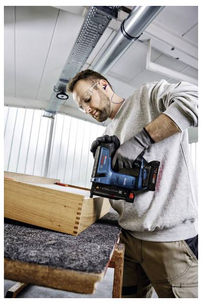 Eine Person mit Sicherheitsbrille und Handschuhen verwendet ein Elektrowerkzeug, um an einem Holzstück in einer Werkstattumgebung zu arbeiten.
