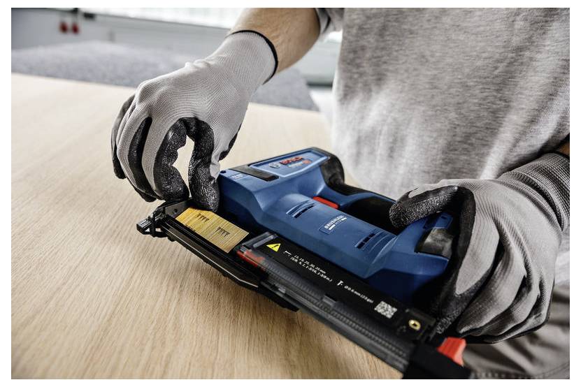 Eine Person mit Handschuhen benutzt ein blaues Elektrowerkzeug auf einer Holzoberfläche und richtet Nägel für Bau- oder Zimmerarbeiten aus.