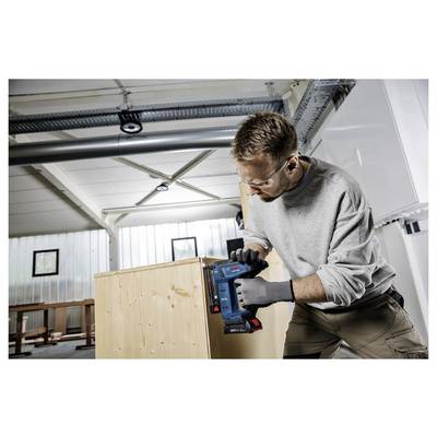 Eine Person mit Sicherheitsbrille und Handschuhen verwendet einen blauen Nageler, um eine Holzkiste in einer Werkstatt zusammenzubauen.