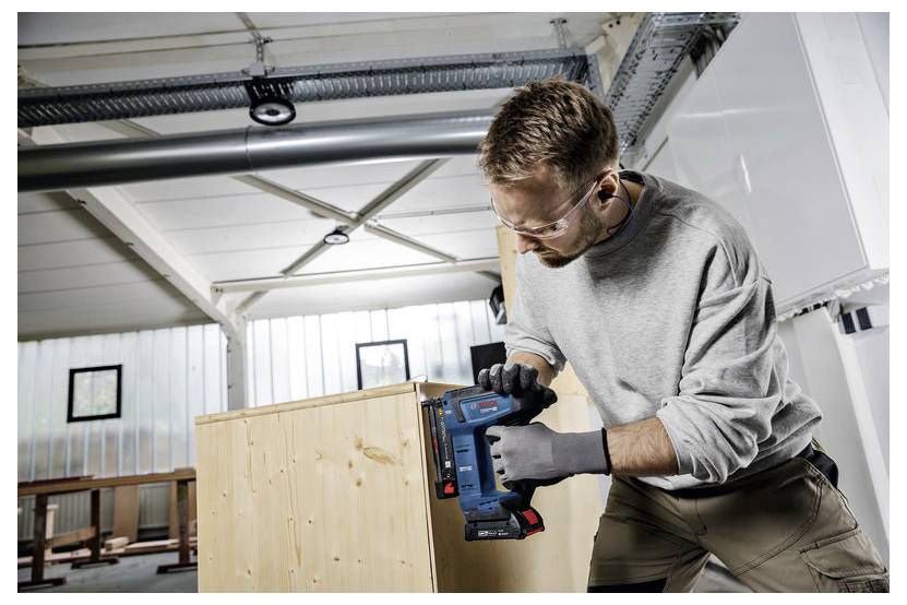 Ein Mann mit Schutzbrille und Handschuhen benutzt ein blaues Elektrowerkzeug, um an einem hellen Holzschrank in einer Werkstatt zu arbeiten.