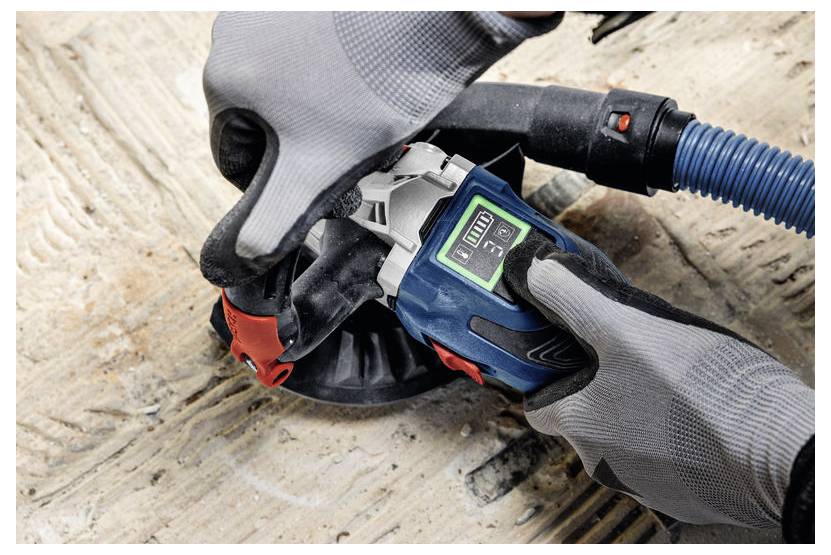 Hände mit Handschuhen bedienen ein tragbares Schleifwerkzeug an Holz, wobei ein digitales Display Einstellungen anzeigt. Ein Staubabsaughose ist angeschlossen.