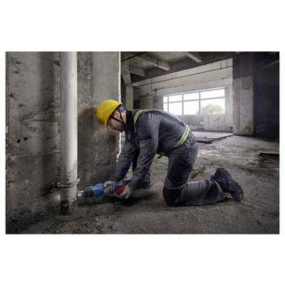 Ein Bauarbeiter in Schutzausrüstung kniet auf einem Betonboden und verwendet ein Elektrowerkzeug, um an einem vertikalen Rohr in einem großen, unfertig wirkenden Raum zu arbeiten.