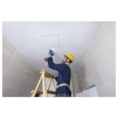 Ein Bauarbeiter mit Schutzhelm und Sicherheitsgurt verwendet einen Bohrer, um ein Quadrat in eine Decke zu schneiden, während er auf einer Holzleiter in einem gekachelten Raum steht.