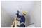 Ein Bauarbeiter mit Schutzhelm und Sicherheitsgurt verwendet einen Bohrer, um ein Quadrat in eine Decke zu schneiden, während er auf einer Holzleiter in einem gekachelten Raum steht.