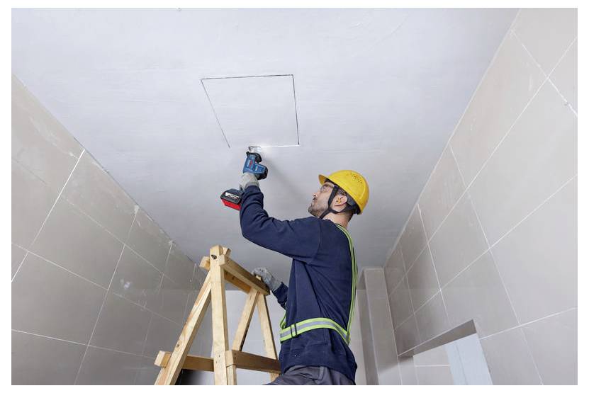 Ein Arbeiter in Sicherheitsausrüstung steht auf einer Leiter und verwendet einen Bohrer an einer Deckenplatte in einem gefliesten Raum, was auf Wartungs- oder Installationsarbeiten hindeutet.