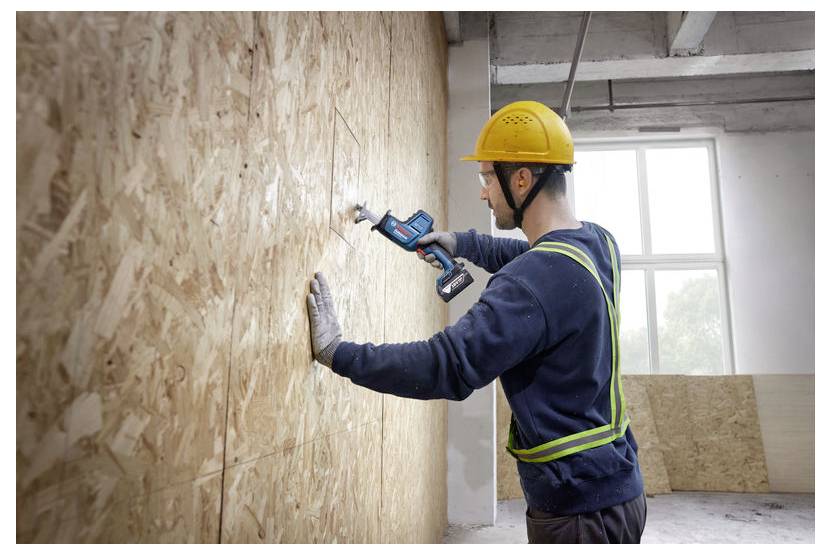 Ein Bauarbeiter mit gelbem Schutzhelm und Sicherheitsgurt bohrt mit einem Akkuschrauber an einer Sperrholzwand in einem im Bau befindlichen Gebäude.