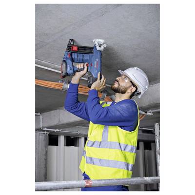 Ein Bauarbeiter mit Schutzhelm und Sicherheitsweste benutzt einen Bohrer, um an einer Decke zu arbeiten, während er auf einem Gerüst in einem im Bau befindlichen Gebäude steht.