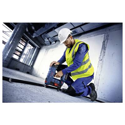 Ein Bauarbeiter in Sicherheitsausrüstung kniet nieder, um ein Elektrowerkzeug auf dem Boden einer Baustelle zu bedienen, und konzentriert sich mit voller Aufmerksamkeit auf seine Aufgabe.