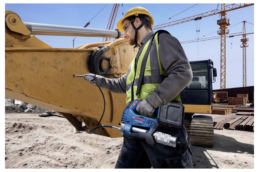 Ein Bauarbeiter mit Schutzhelm und Warnweste bedient ein Elektrowerkzeug an schwerer Baumaschine auf einer Baustelle mit Kränen im Hintergrund.