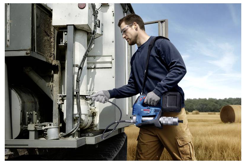 Eine Person bedient ein blaues und schwarzes elektrisches Werkzeug neben einer großen Industriemaschine auf einem Feld mit Heuballen im Hintergrund.