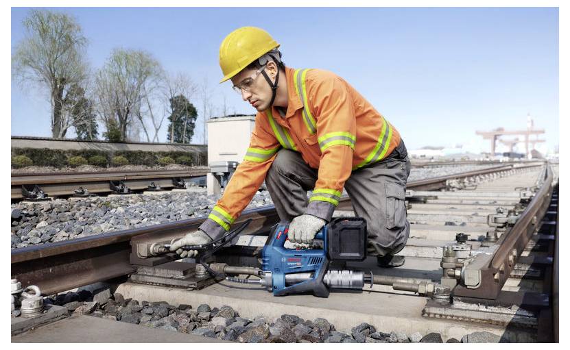 Ein Arbeiter in einer orangefarbenen Jacke und einem gelben Helm benutzt ein blaues Elektrowerkzeug zur Wartung von Eisenbahnschienen an einem klaren Tag.