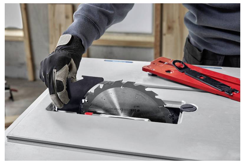 Eine Person trägt einen Handschuh und stellt eine Kreissägeblatt an einer Tischkreissäge ein, wobei Werkzeuge auf dem Tisch in einer Werkstattumgebung sichtbar sind.