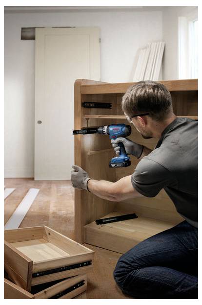 Eine Person montiert Holzmöbel mit einer Bohrmaschine, wobei Komponenten und Werkzeuge auf einem Hartholzboden verstreut liegen.