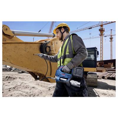 Ein Bauarbeiter mit Sicherheitsweste und Schutzhelm bedient ein Werkzeug an einem Bagger auf einer geschäftigen Baustelle mit Kränen im Hintergrund.