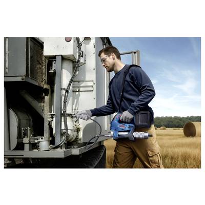 Eine Person verwendet ein blaues Werkzeug, um eine Maschine auf einem großen Fahrzeug in einem ländlichen Feld zu reparieren oder zu warten, wobei im Hintergrund Heuballen zu sehen sind.