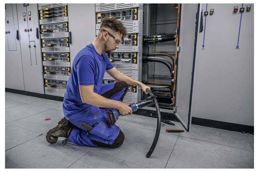 Eine Person mit Sicherheitsbrille und blauer Arbeitskleidung kniet und benutzt eine Bohrmaschine oder ein ähnliches Werkzeug an einem großen Kabel in einer industriellen Umgebung.