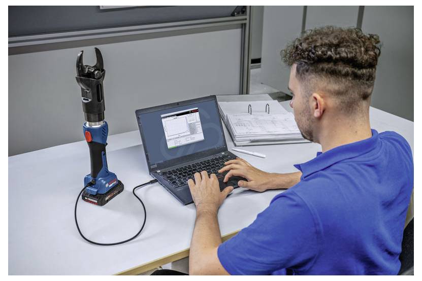 Eine Person in einem blauen Hemd arbeitet an einem Laptop, der mit einem kabellosen Crimpwerkzeug verbunden ist, während technische Dokumente daneben geöffnet sind.