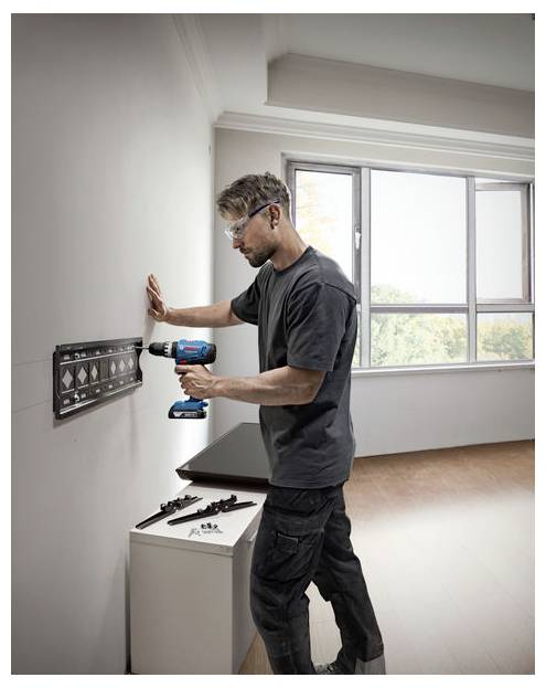 Eine Person mit Sicherheitsbrille verwendet einen Bohrer, um eine TV-Halterung an einer Wand in einem hellen, modernen Raum mit einem großen Fenster zu installieren.