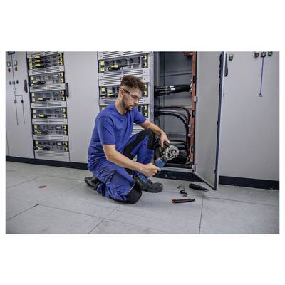 Ein Techniker in blauer Arbeitskleidung kniet auf einem Fliesenboden und bedient ein Handwerkzeug vor einem offenen Elektroschrank mit verschiedenen Kabeln.
