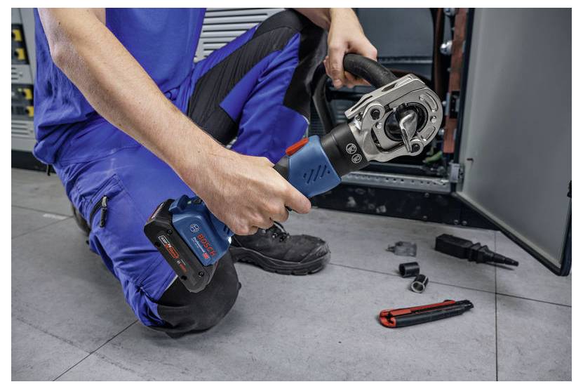 Eine Person in blauer Arbeitskleidung kniet auf dem Boden und verwendet ein Elektrowerkzeug, um eine Komponente einer elektrischen Schalttafel zu reparieren oder einzustellen. Werkzeuge befinden sich in der Nähe.