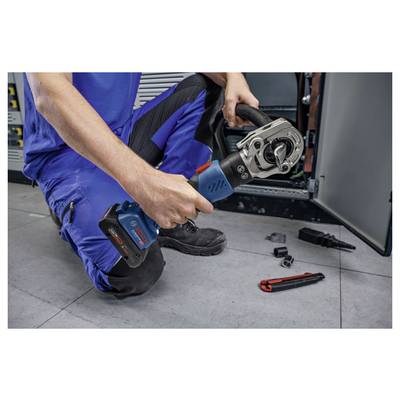 Eine Person in blauer Arbeitskleidung kniet auf dem Boden und verwendet ein Elektrowerkzeug, um eine Komponente einer elektrischen Schalttafel zu reparieren oder einzustellen. Werkzeuge befinden sich in der Nähe.