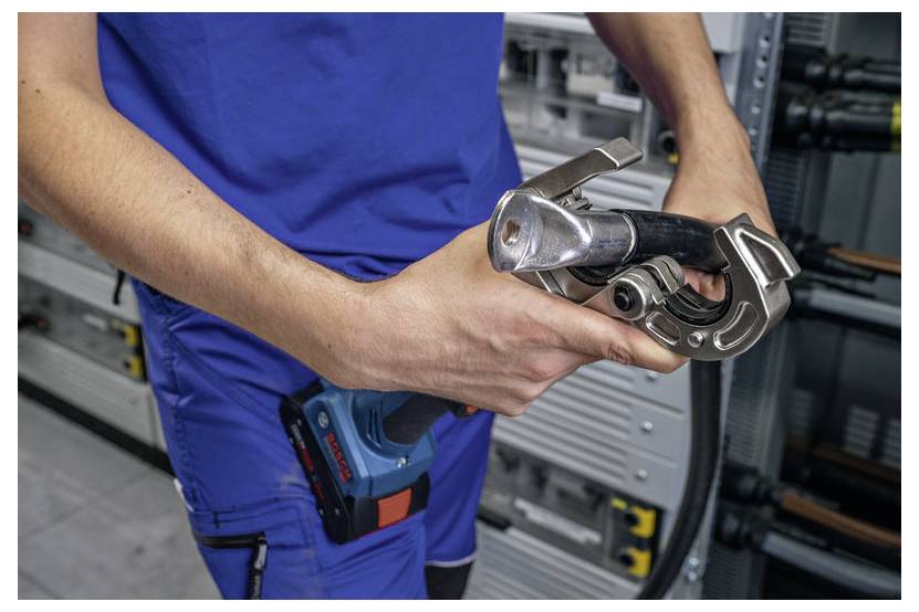 „Person in blauer Uniform, die ein Crimpwerkzeug in der Nähe von Elektrotafeln hält und sich darauf vorbereitet, Leitungen zu verbinden oder anzupassen."