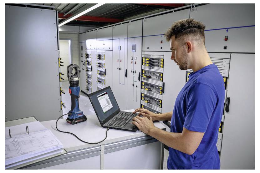 Eine Person in einem blauen Hemd bedient einen Laptop in einem elektrischen Kontrollraum, umgeben von Bedienfeldern und Ausrüstung für Wartungsaufgaben.