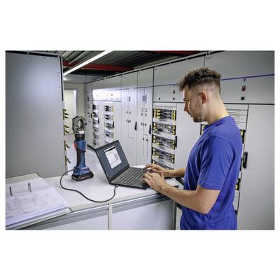 Eine Person in einem blauen Hemd bedient einen Laptop in einem elektrischen Kontrollraum, umgeben von Bedienfeldern und Ausrüstung für Wartungsaufgaben.