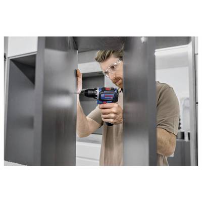 Eine Person benutzt einen blauen Akkuschrauber, um ein Loch in einen Metallschrank zu bohren. Sie trägt durchsichtige Schutzbrille und ein beiges Hemd.