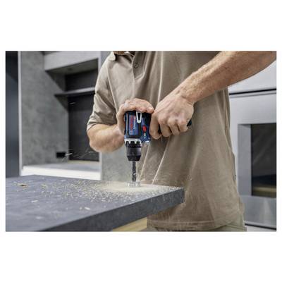 Eine Person benutzt eine Bohrmaschine, um ein Loch in eine Arbeitsplatte zu bohren, während Holzspäne in der Umgebung verstreut werden. Die Umgebung scheint eine Werkstatt oder eine Küche zu sein.