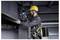 Ein Bauarbeiter in Sicherheitsausrüstung, einschließlich Helm und Sicherheitsgurt, verwendet ein Elektrowerkzeug, um Metallträger in einer industriellen Umgebung zu befestigen.