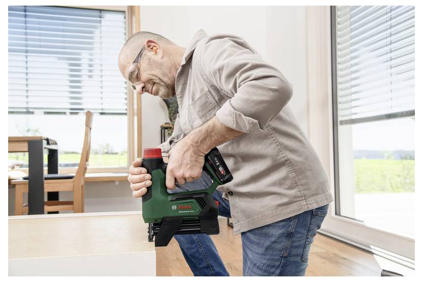 Eine Person mit Sicherheitsbrille verwendet ein grünes Werkzeug zum Möbelbau in einem hell erleuchteten Raum mit großen Fenstern.