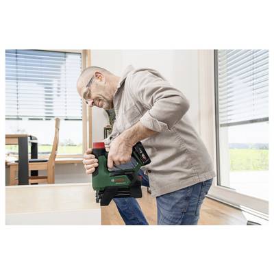 Eine Person mit Sicherheitsbrille verwendet ein grünes Werkzeug zum Möbelbau in einem hell erleuchteten Raum mit großen Fenstern.