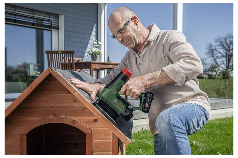 Eine Person verwendet ein Elektrowerkzeug, um Dacheindeckungsmaterial an einer kleinen Holzkonstruktion in einem Garten zu befestigen, wobei Gartenmöbel im Hintergrund zu sehen sind.