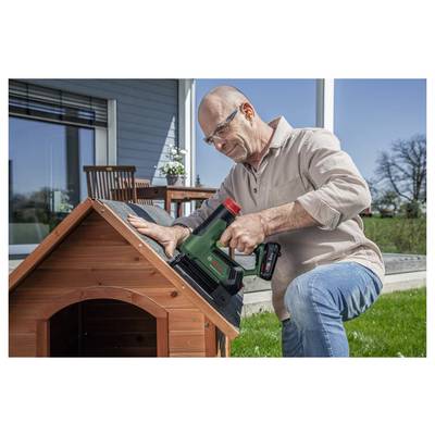 Eine Person verwendet ein Elektrowerkzeug, um Dacheindeckungsmaterial an einer kleinen Holzkonstruktion in einem Garten zu befestigen, wobei Gartenmöbel im Hintergrund zu sehen sind.