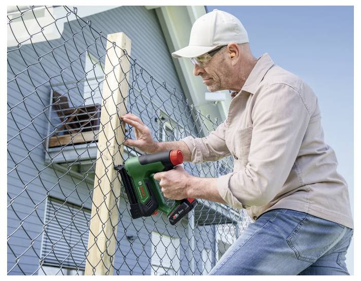 Mann, der einen kabellosen Nagler an einem Holzpfosten, der an einem Maschendrahtzaun befestigt ist, verwendet, trägt Schutzbrille und eine Kappe, mit einem Haus im Hintergrund.
