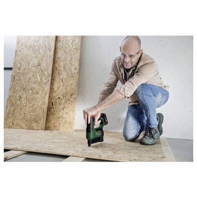 Mann, der ein grünes Elektrowerkzeug auf Holz verwendet, kniend mit Schutzbrille, was auf Zimmermannsarbeit oder Bauaktivität hindeutet.