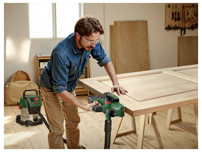 Eine Person mit Schutzbrille, die ein grünes Vakuumwerkzeug auf einem Holztisch in einer Werkstatt verwendet, mit Holzpaneelen und Werkzeugen im Hintergrund.