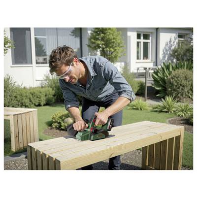 Eine Person mit Sicherheitsbrille benutzt ein Elektrowerkzeug, um eine Holzbank im Freien zu hobeln, umgeben von Pflanzen und einem Gebäude im Hintergrund.