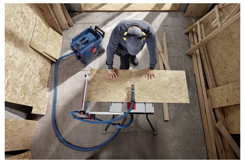 Eine Person mit Mütze und Kopfhörern steht über einer Werkbank und misst ein Holzbrett mit einem Maßband.