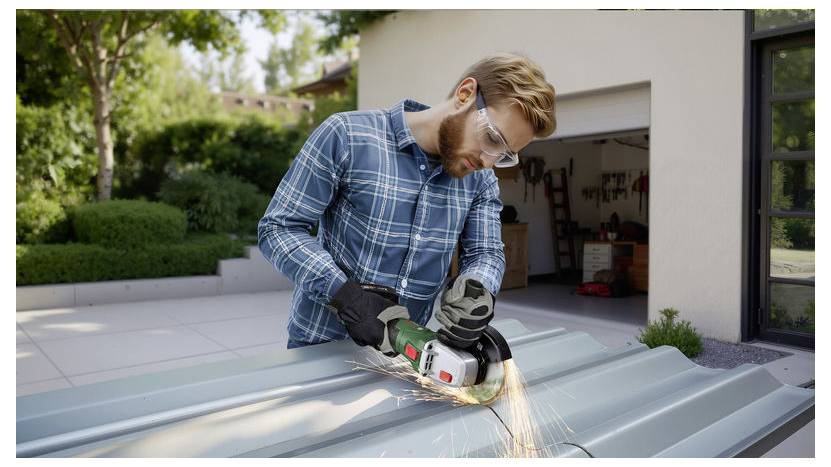 Ein Mann mit Sicherheitsschutzbrille verwendet ein grün-schwarzes Elektrowerkzeug, um Metallbleche außerhalb einer Garage in einem Wohngebiet zu schneiden.
