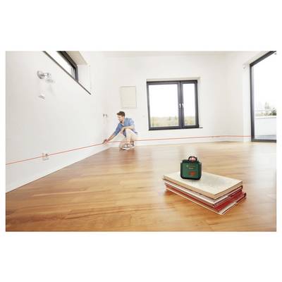 Eine Person vermisst einen Raum mit einem Lasernivelliergerät und legt eine rote Laserlinie entlang einer weißen Wand. Ein Stapel Bücher befindet sich auf dem Holzboden.