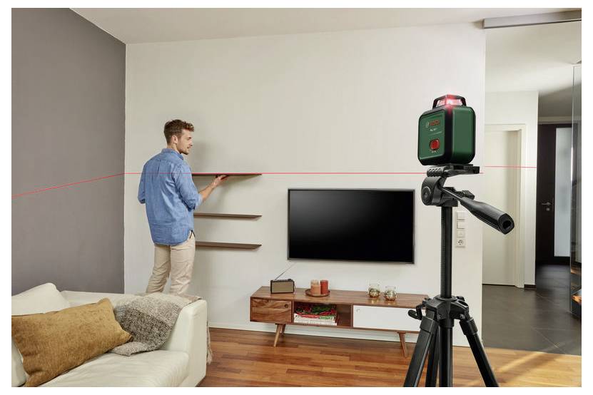 Eine Person verwendet eine Laserwasserwaage, um ein Regal an einer weißen Wand in einem modernen Wohnzimmer mit einer Couch und einem wandmontierten Fernseher auszurichten.