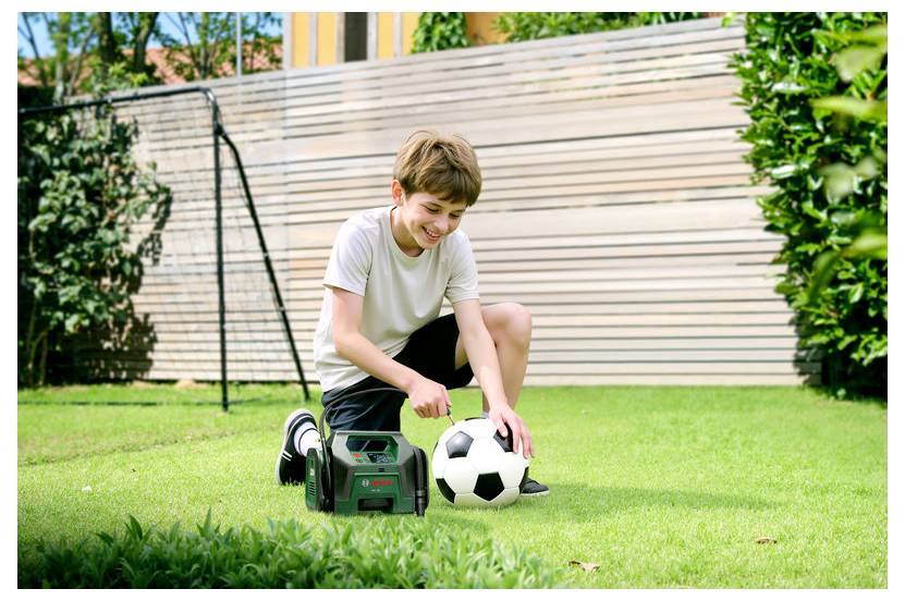 Ein Junge kniet auf einer grasbewachsenen Wiese und lächelt, während er mit einem schwarz-weißen Fußball interagiert. Um ihn herum befinden sich grüne Pflanzen und ein Zaun.