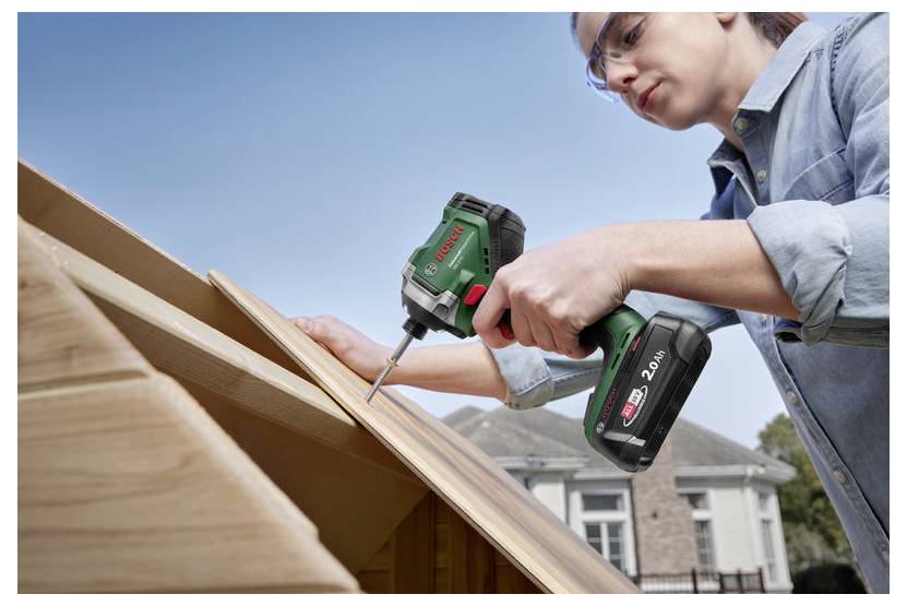 Eine Person, die einen kabellosen Akkuschrauber verwendet, um eine Holzplatte an einer Konstruktion zu befestigen, mit einem Haus im Hintergrund unter einem klaren Himmel.