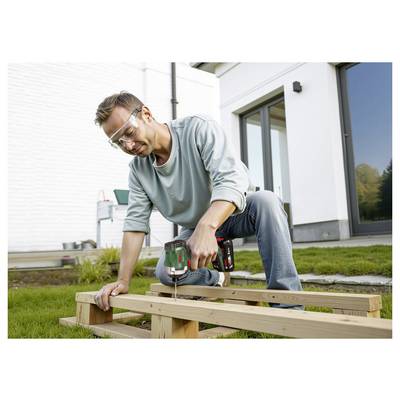 Eine Person kniet in einem Hinterhof, trägt Schutzbrille und verwendet eine Bohrmaschine an Holzplanken, um eine Struktur außerhalb eines modernen Hauses zu bauen.