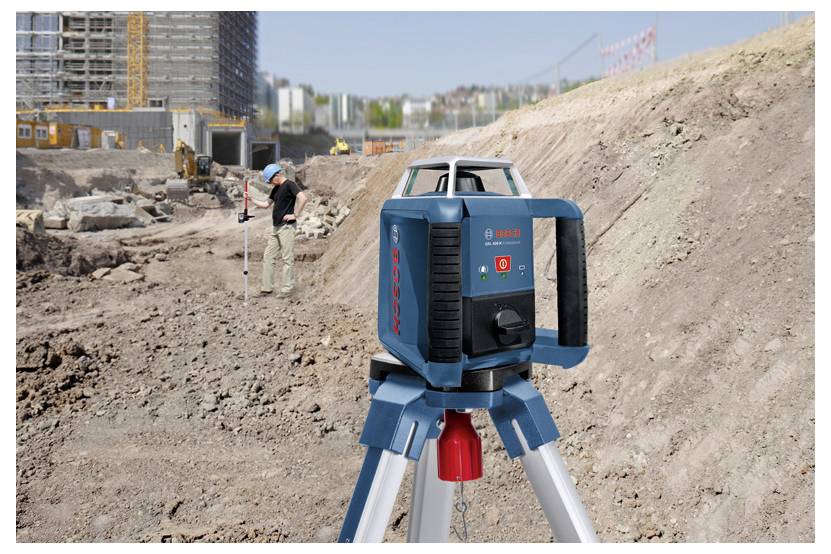Ein Lasernivelliergerät auf einem Stativ ist auf einer Baustelle aufgestellt, während im Hintergrund ein Arbeiter die Höhe misst.